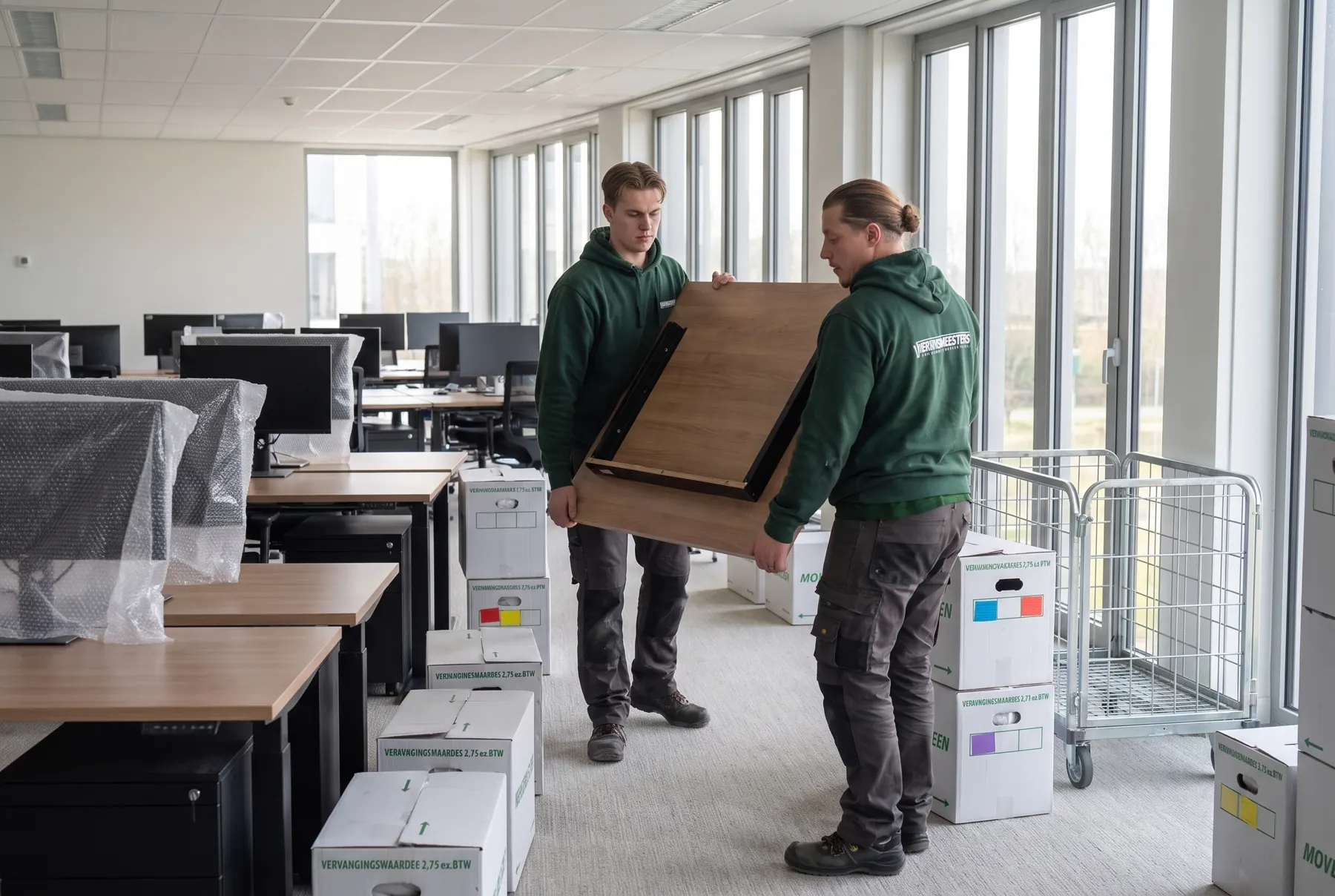 Twee verhuizers dragen een bureaublad door een leeg kantoor met ingepakte beeldschermen en verhuisdozen